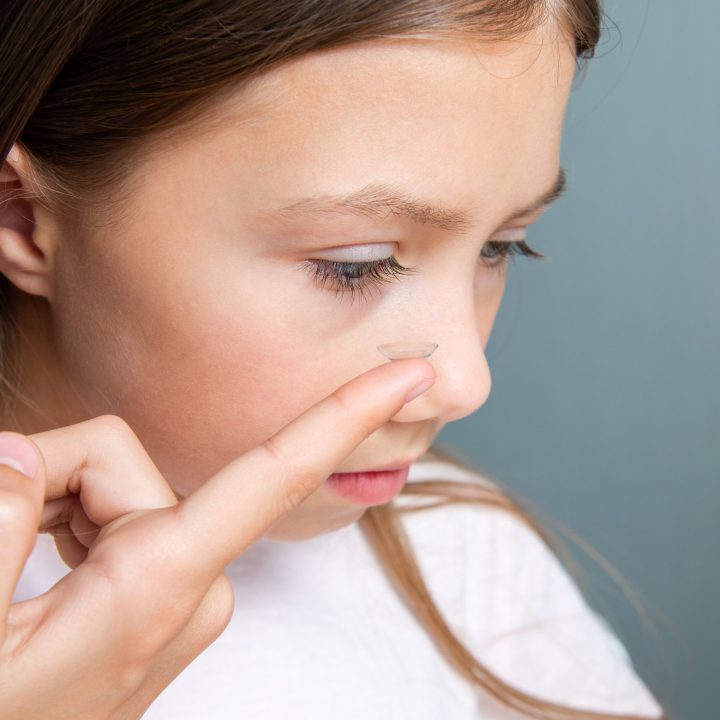 Little child girl inserting a transparent contact lens into her eye on gray background. The concept of ophthalmological poor sight correction