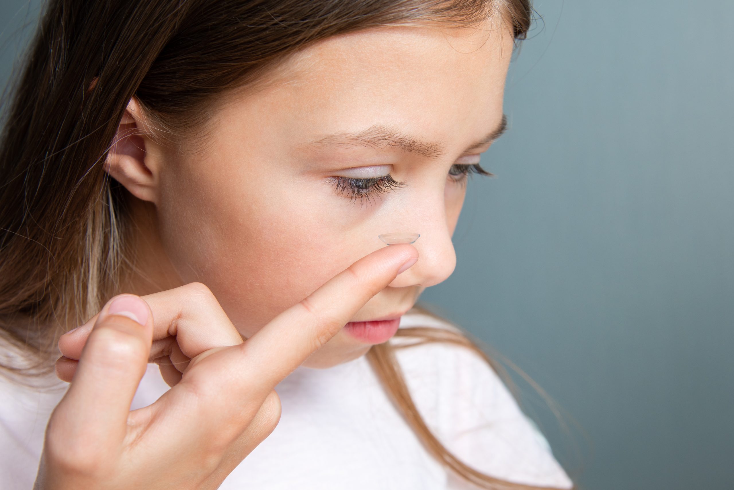 Little child girl inserting a transparent contact lens into her eye on gray background. The concept of ophthalmological poor sight correction