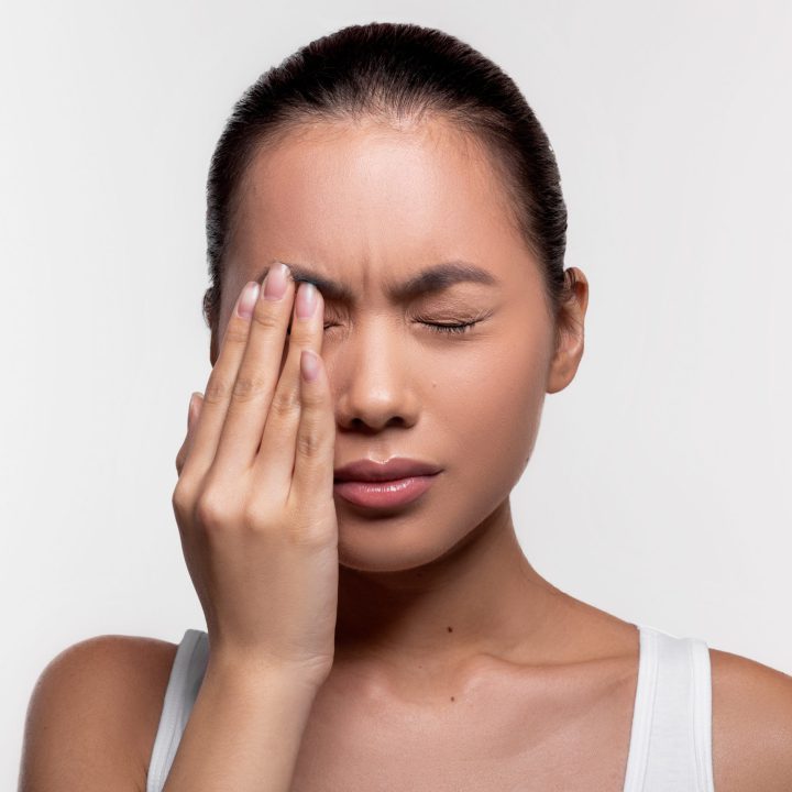 Upset young asian woman suffering from strong eye pain, touching her eye and crying over white studio background, closeup photo, copy space. Healthcare, eye disorders and diseases concept